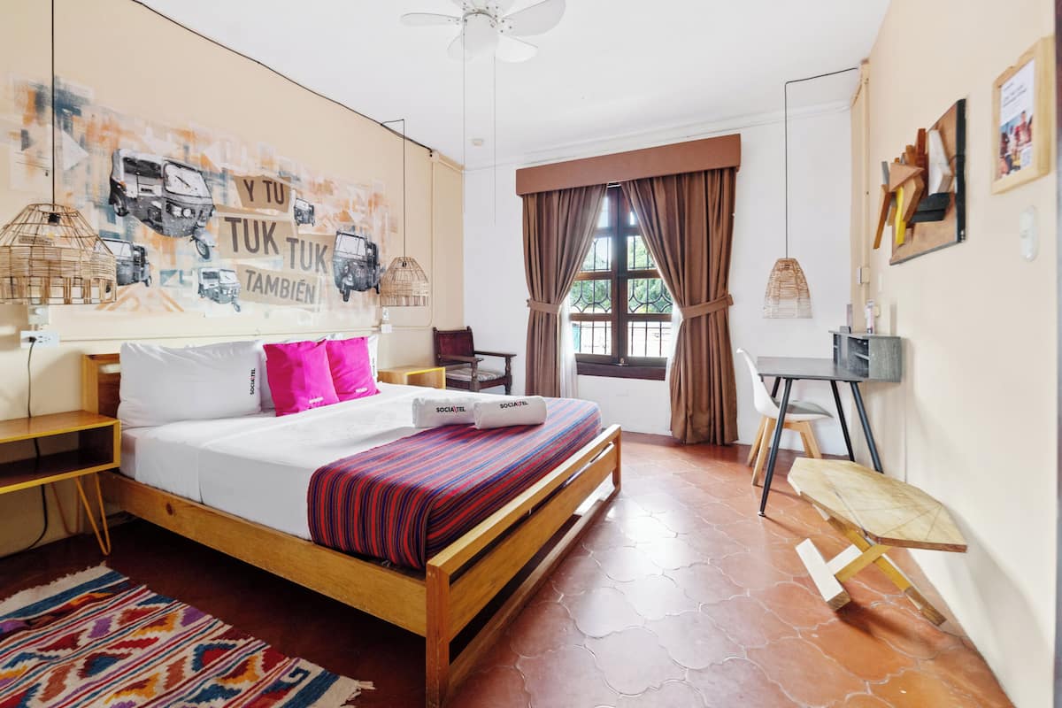 Une chambre d’hôtel comprenant un lit, un bureau, une chaise, une fenêtre avec des rideaux et un mur orné d’une fresque.