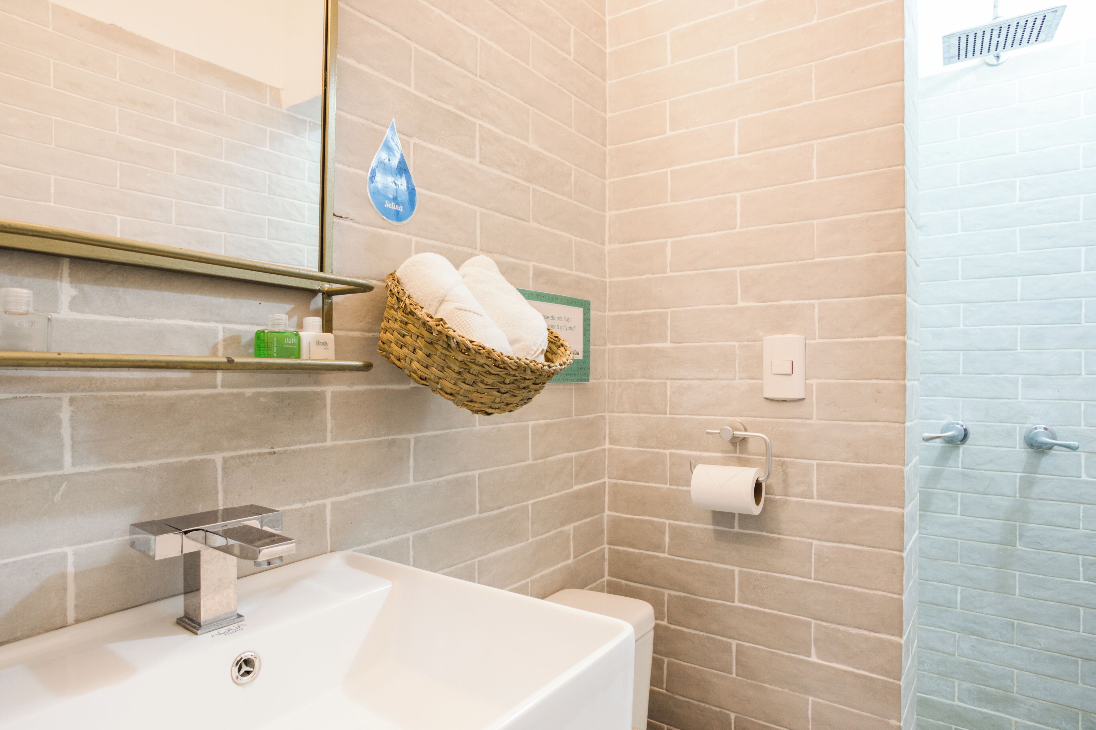 A bathroom with a white sink, a mirror, a shower, and a wall-mounted basket with towels.