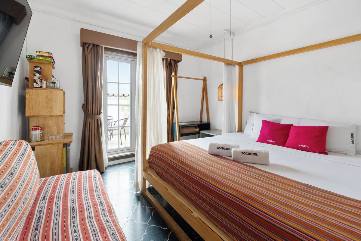 Une chambre d’hôtel avec un lit, deux tables de chevet, une étagère en bois avec des livres et des verres, une fenêtre avec des rideaux et un luminaire suspendu.