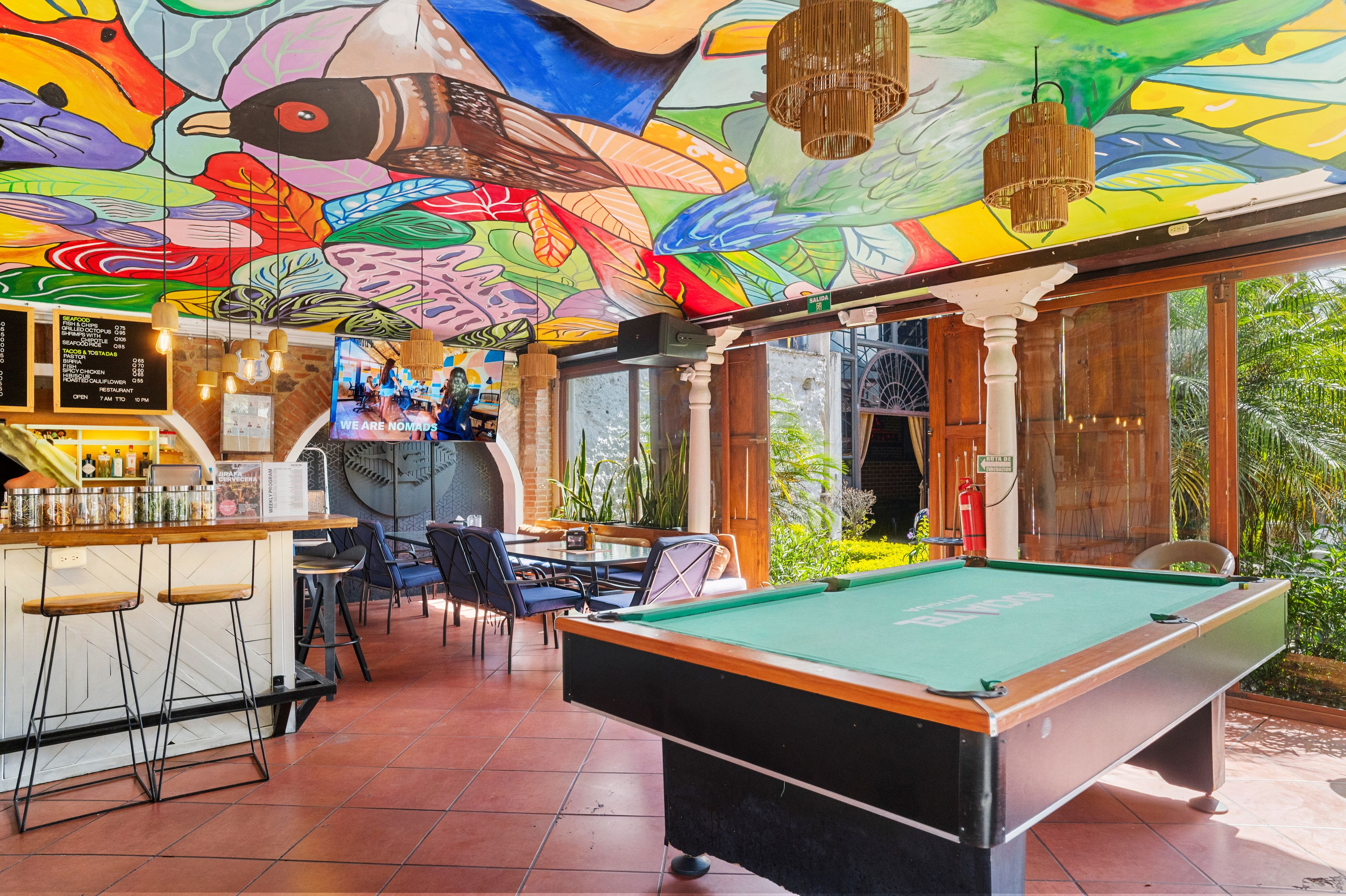 Une table de billard dans une pièce dont le plafond est orné d’une fresque colorée, un coin bar avec des tabourets et une vue sur la verdure à l’extérieur.