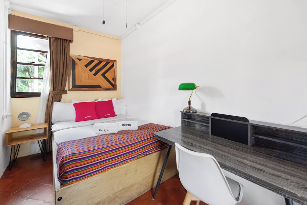 Une petite pièce comprenant un lit, un bureau, une chaise, une fenêtre avec des rideaux et une lampe fixée au mur.