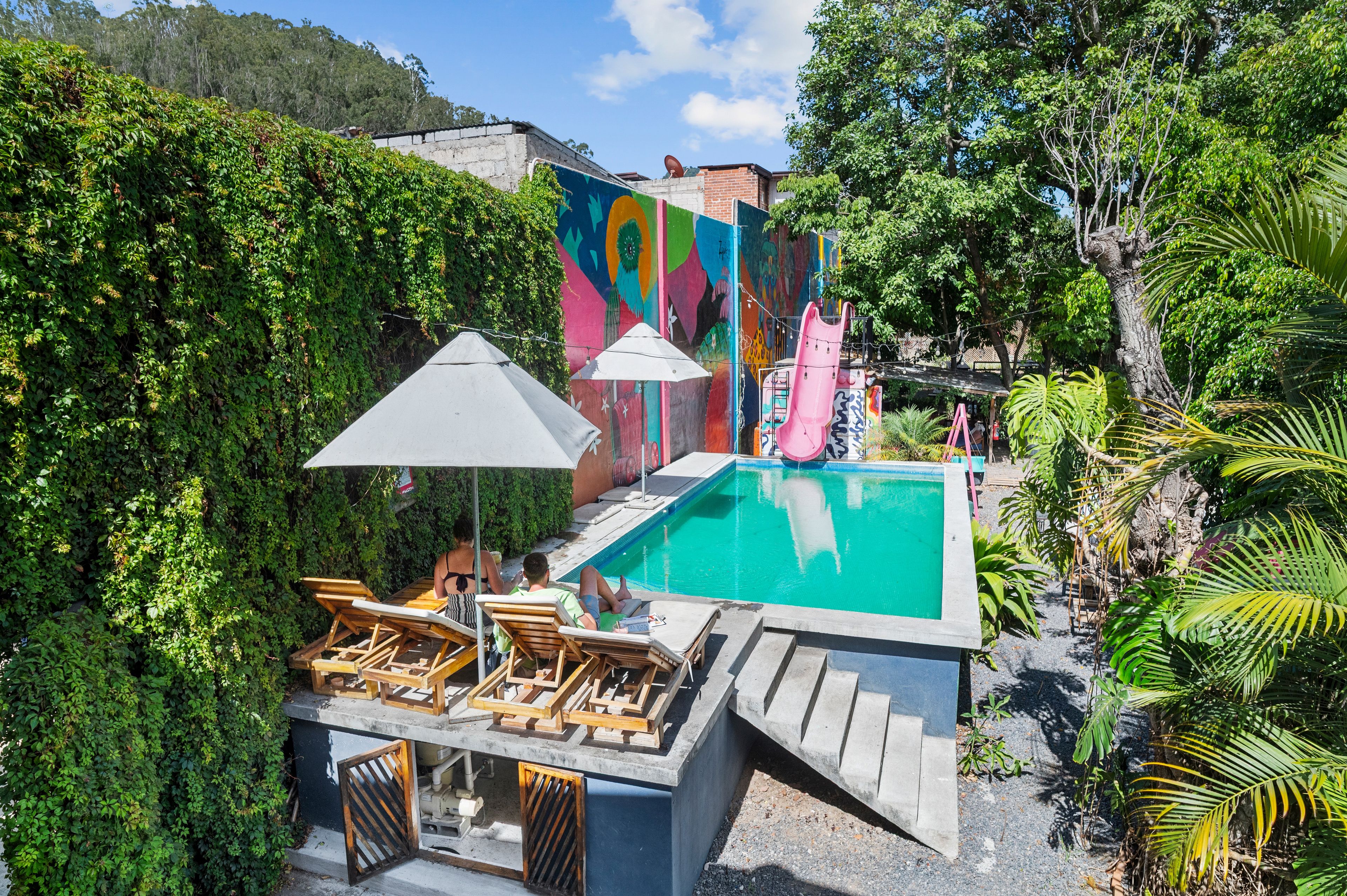 A pool area with lounge chairs and umbrellas, surrounded by lush greenery and a colorful mural.