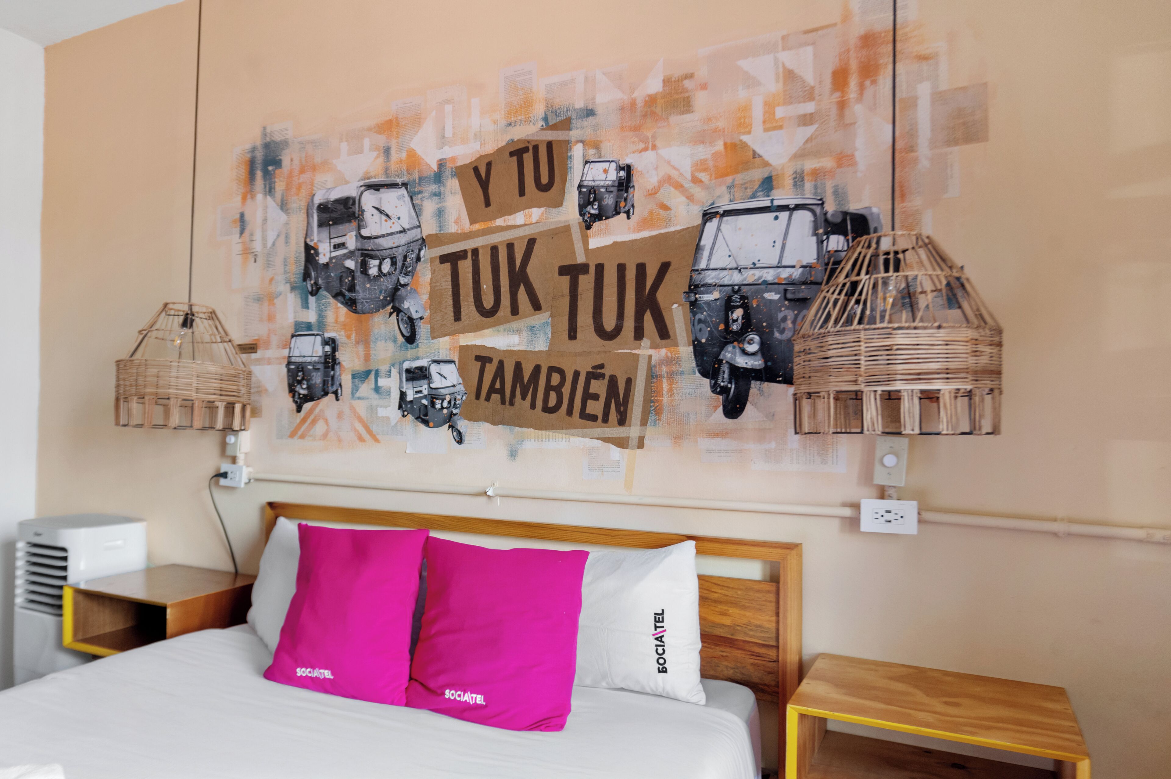 A hotel room with a bed, two pillows, a wall art of vehicles, and a wicker lamp.