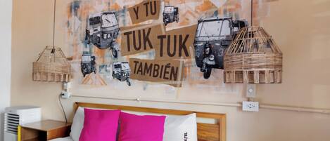 A hotel room with a bed, two pillows, a wall art of vehicles, and a wicker lamp.