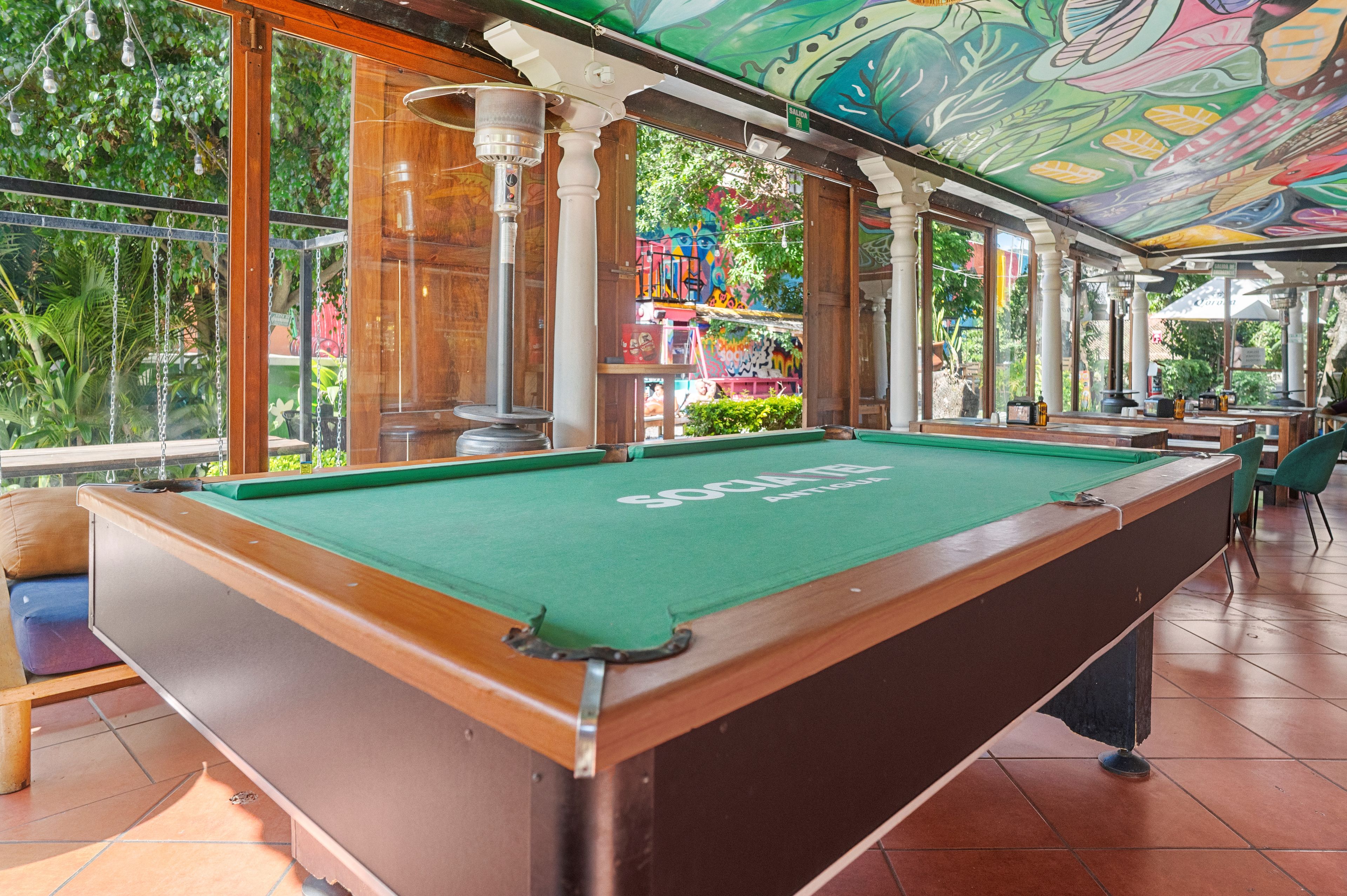 A pool table with a green felt surface, surrounded by wooden furniture and a ceiling with a colorful mural.
