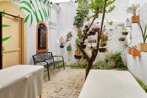 Une cour avec un banc, une table et divers plantes en pot, le tout adossé à un mur blanc.