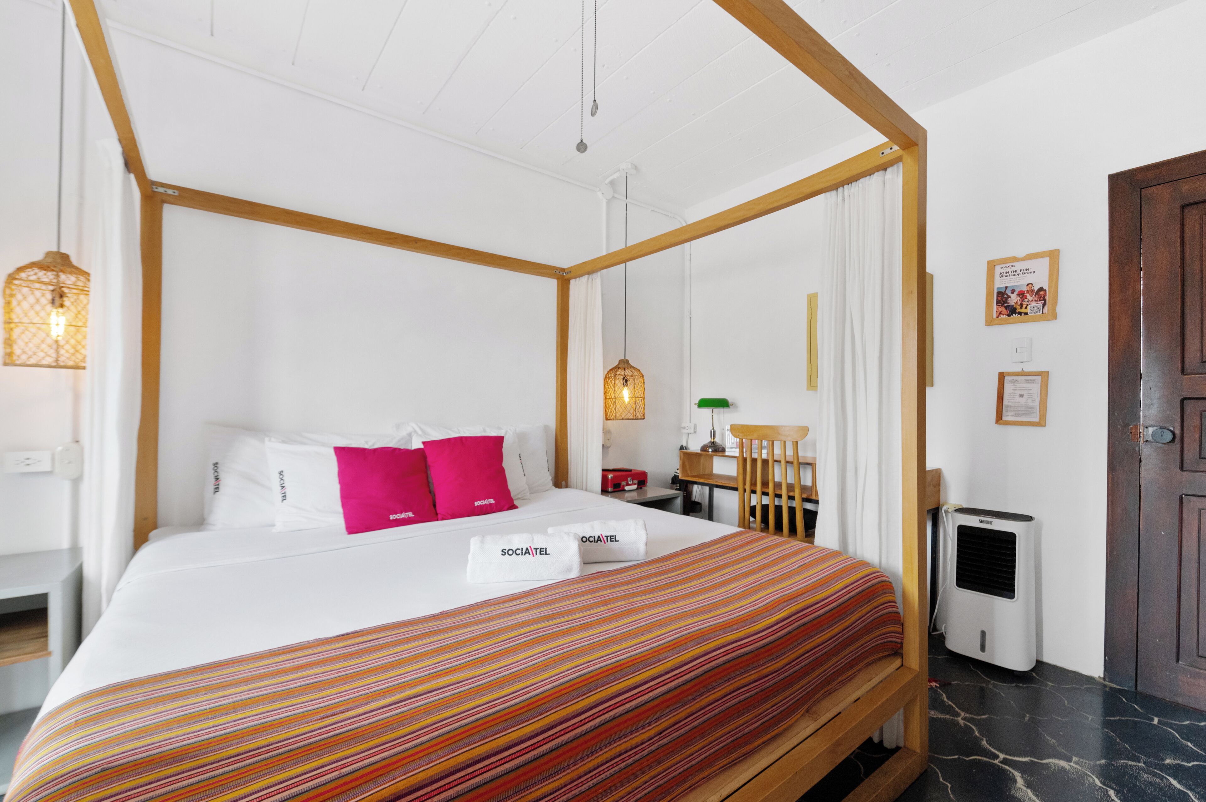 Una cama con dosel tapizada con sábanas blancas y almohadas rosas, un edredón a rayas, un cabecero de madera, una mesita de noche con una lámpara y un aire acondicionado instalado en la pared.
