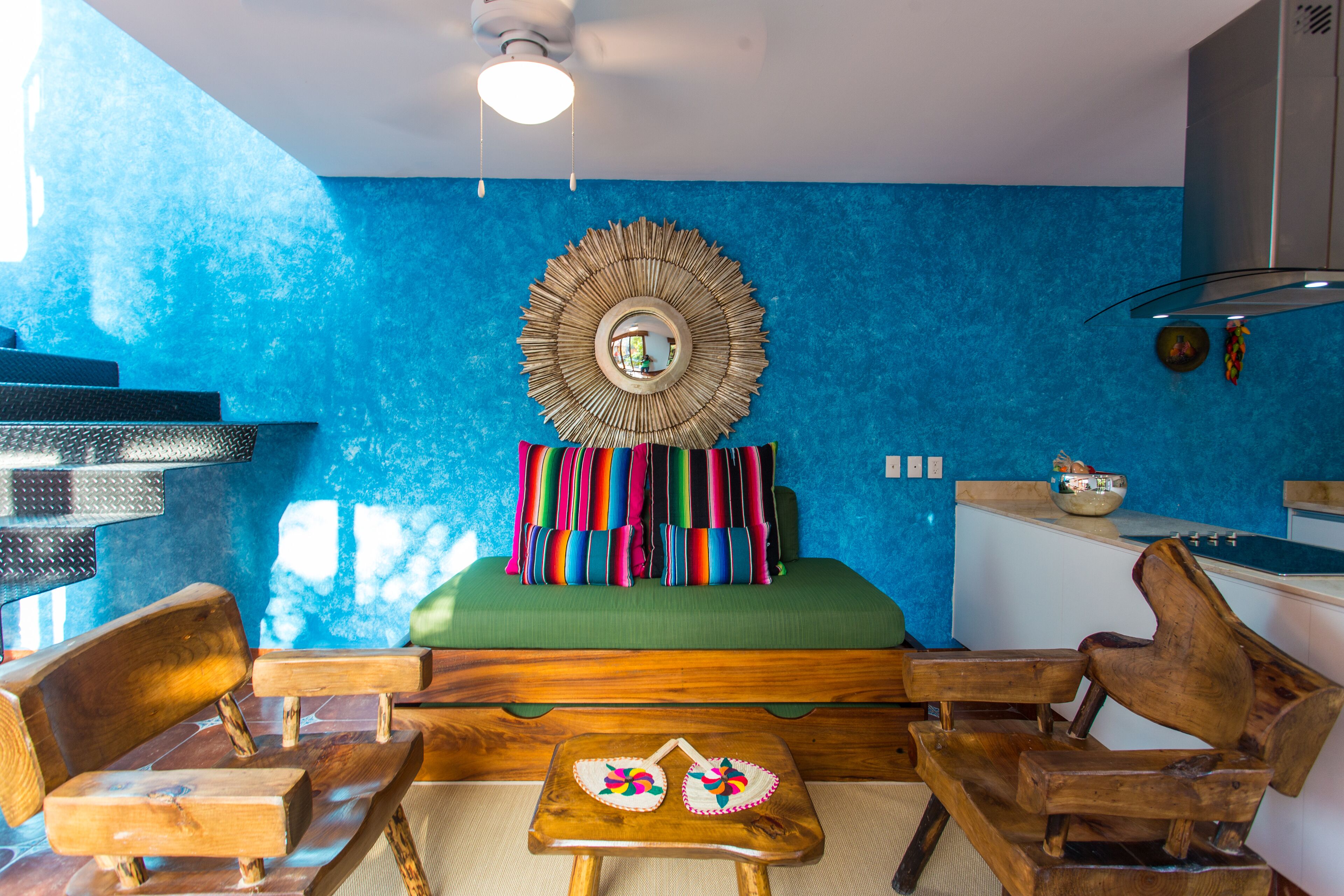 A room with a blue textured wall, a round mirror, a green sofa with colorful pillows, wooden chairs, and a small table with decorative items.