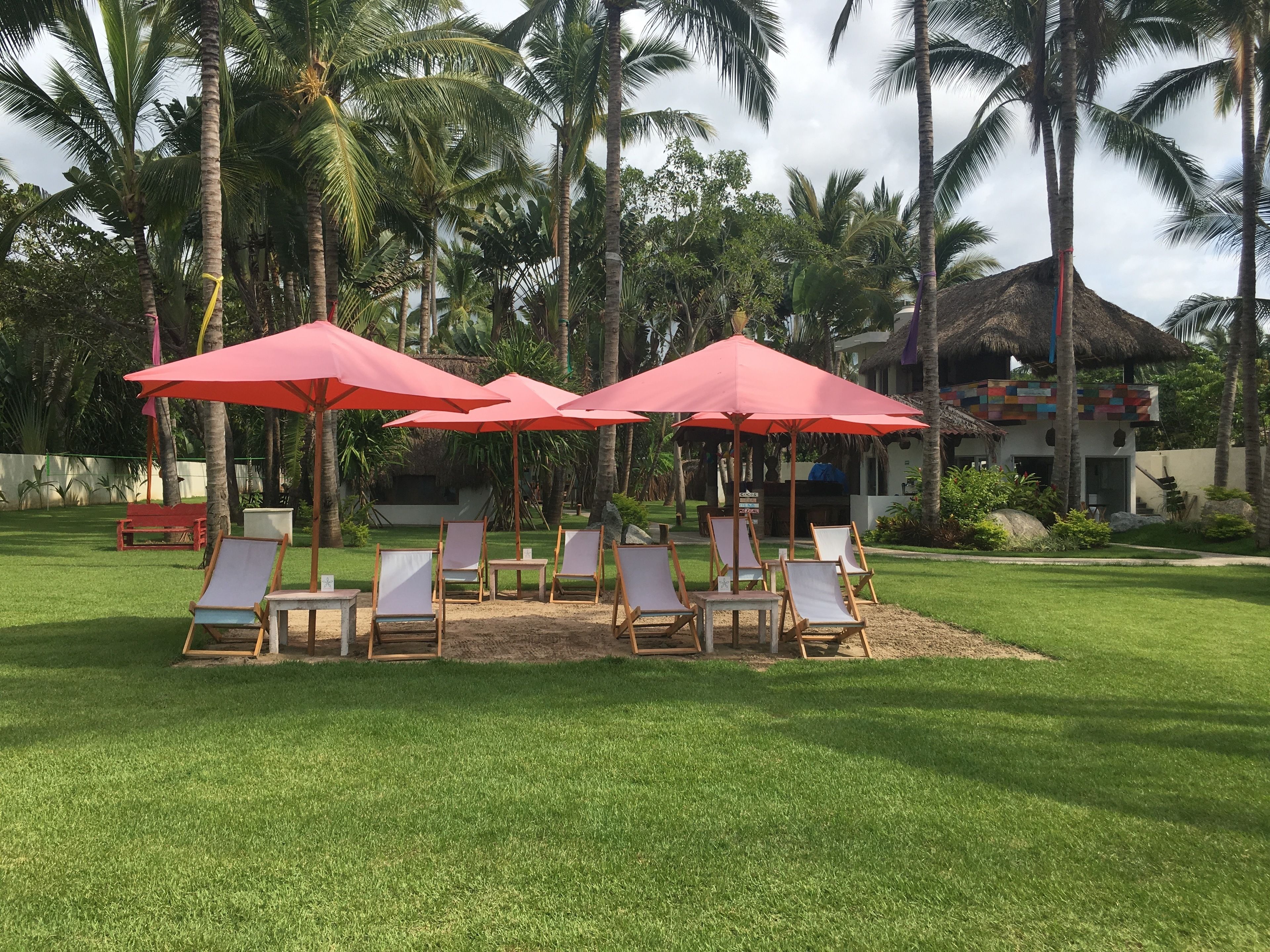 A tropical resort with a lawn area featuring red umbrellas and lounge chairs.