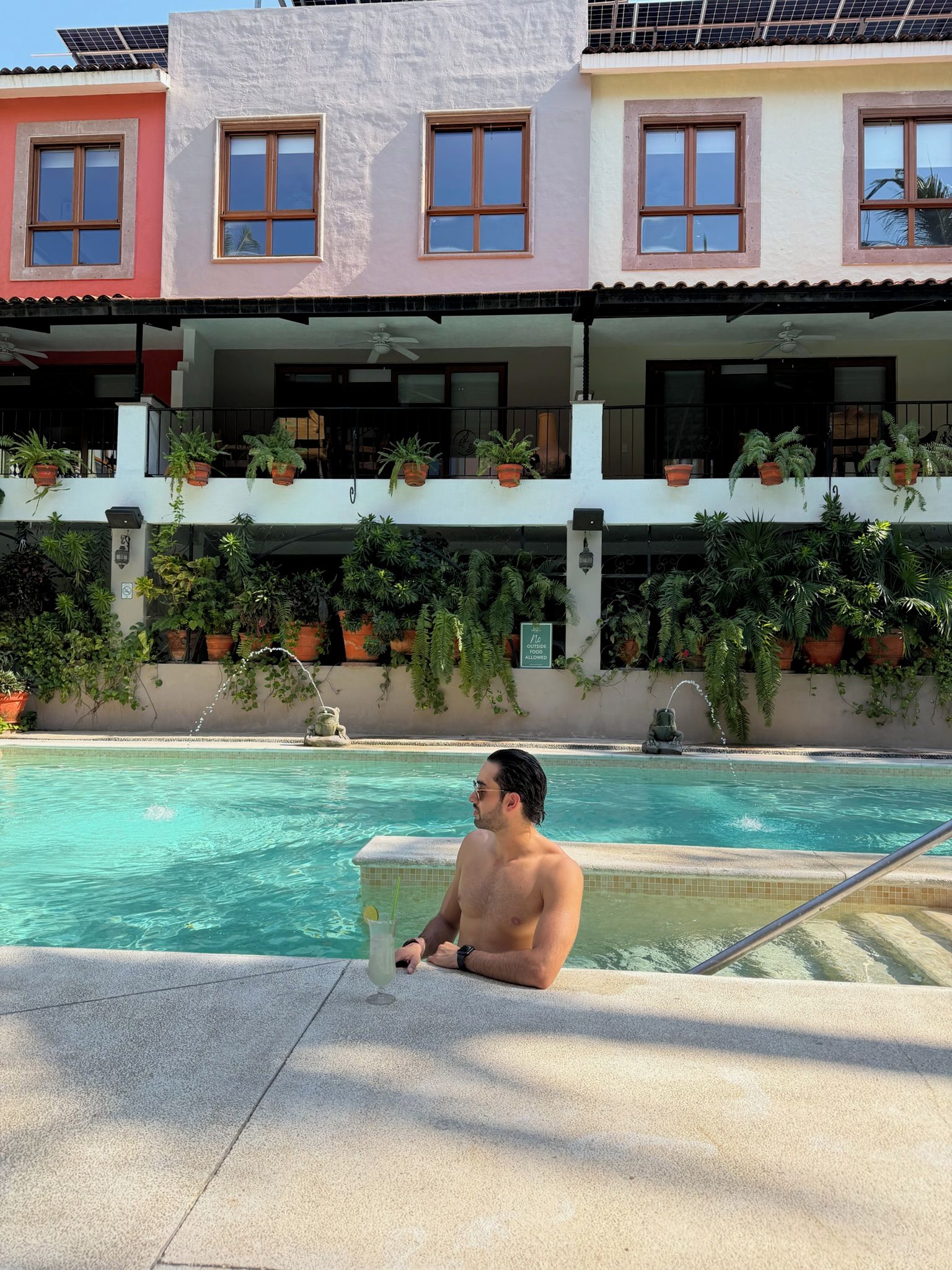 A man sitting by a pool with a clear view of a building with multiple balconies and potted plants.