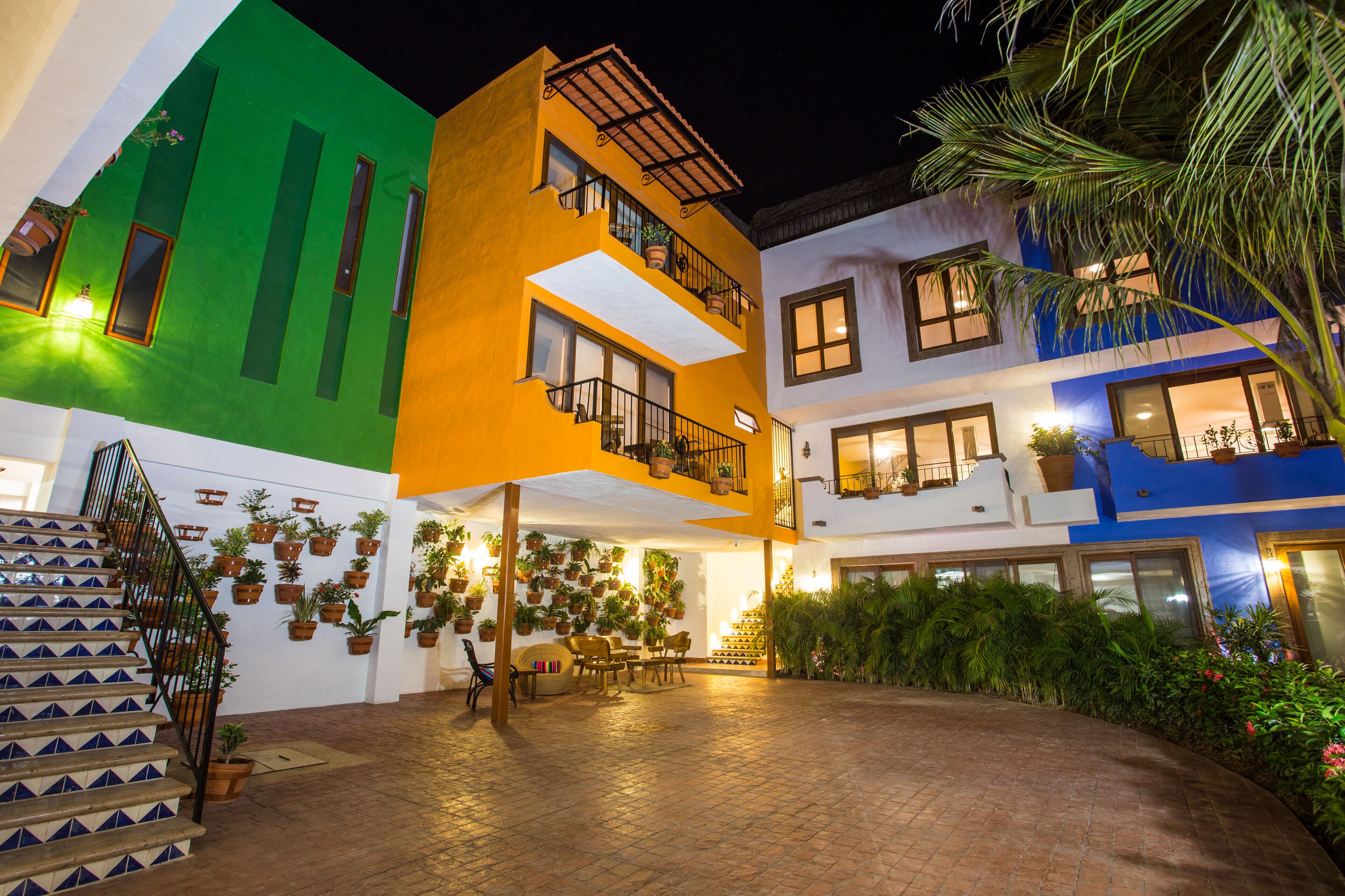 A multi-story building with a green, orange, and blue facade, a covered outdoor seating area with tables and chairs, and a staircase with blue and white tiles.
