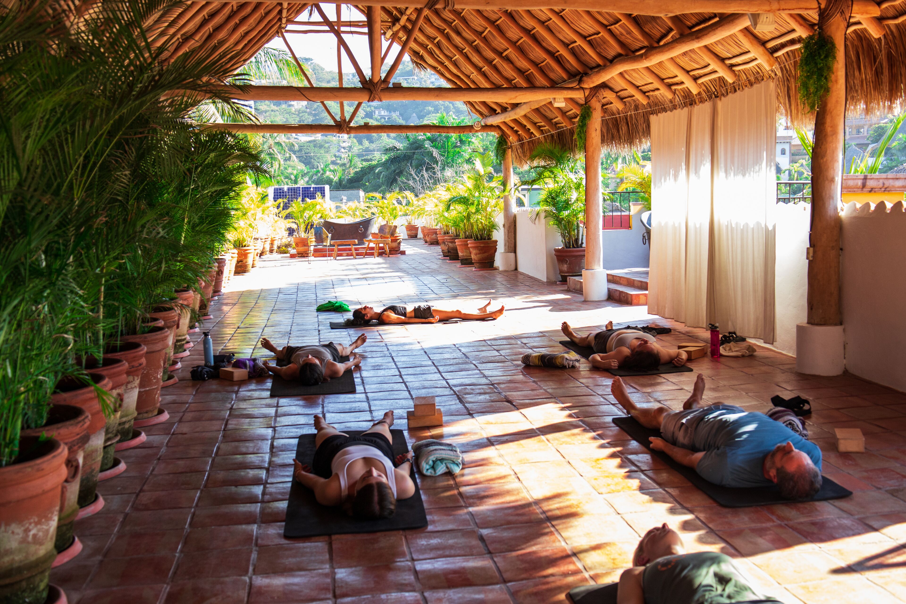 Un espace extérieur abrité où des personnes sont allongées sur des tapis, entourées de plantes en pot, et surmonté d’une structure à toit de chaume.