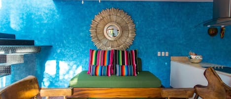 Una habitación con pared azul con textura, un espejo redondo, un banco acolchado verde y sillas de madera.