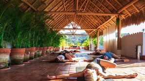 Una clase de yoga en un espacio cubierto con techo de paja, rodeado de plantas en macetas y vistas a la vegetación.
