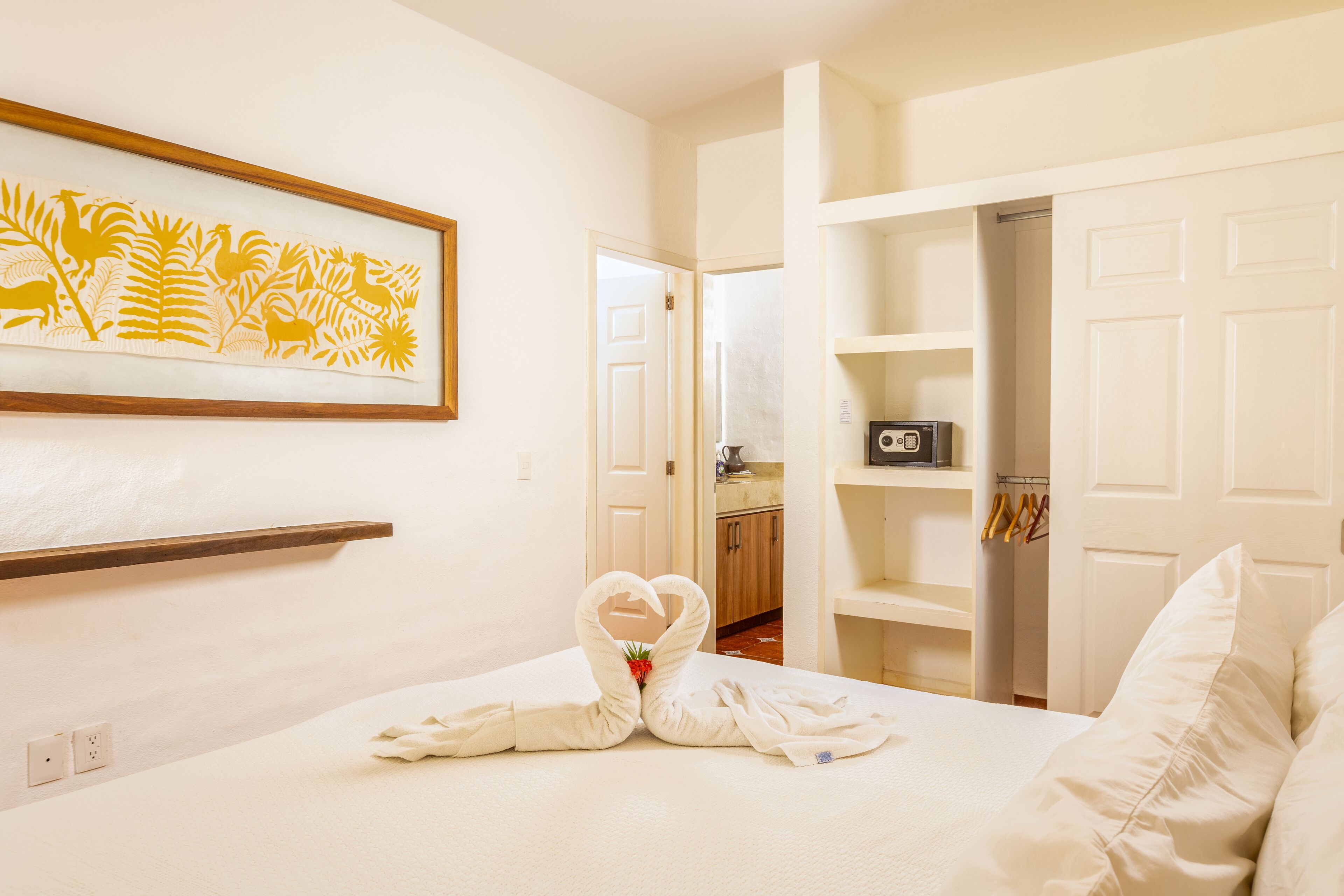 A hotel room with a bed, a heart-shaped towel arrangement, a framed artwork, and a closet with hanging clothes.