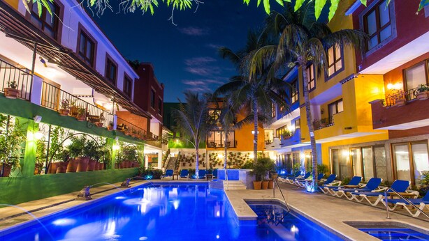 Una piscina con sillas de descanso y plantas en macetas, rodeada de edificios de varios pisos con balcones e iluminación exterior.
