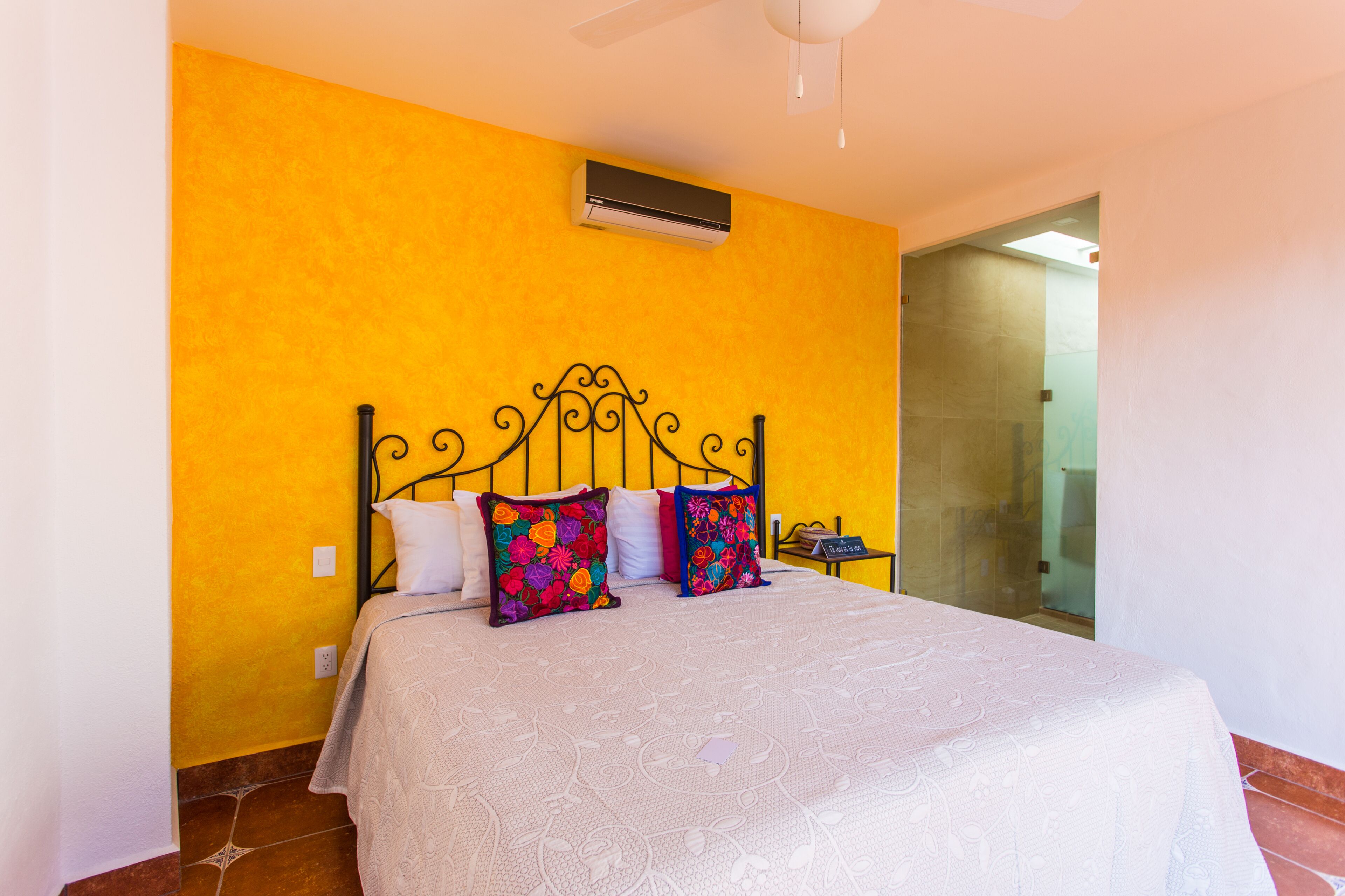 Ein Bett mit Metallgestell, weißer Bettdecke und bunten Kissen vor einer gelben Wand.
