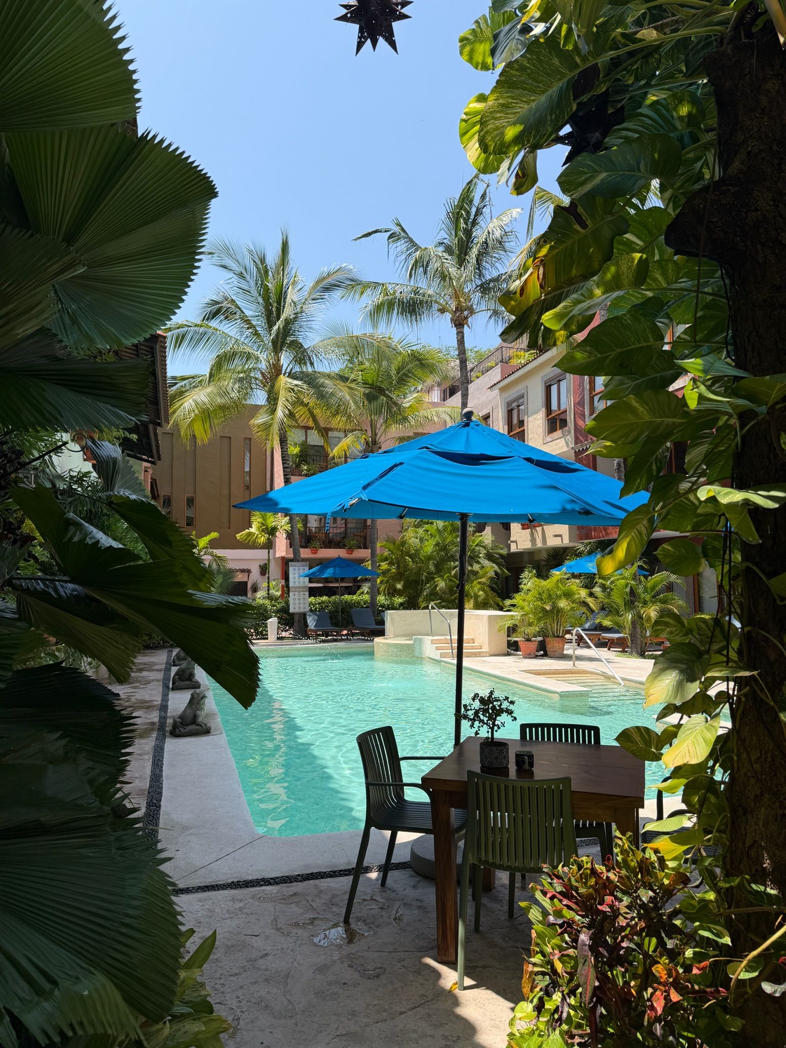A pool area with a table and chairs, surrounded by tropical plants and palm trees.