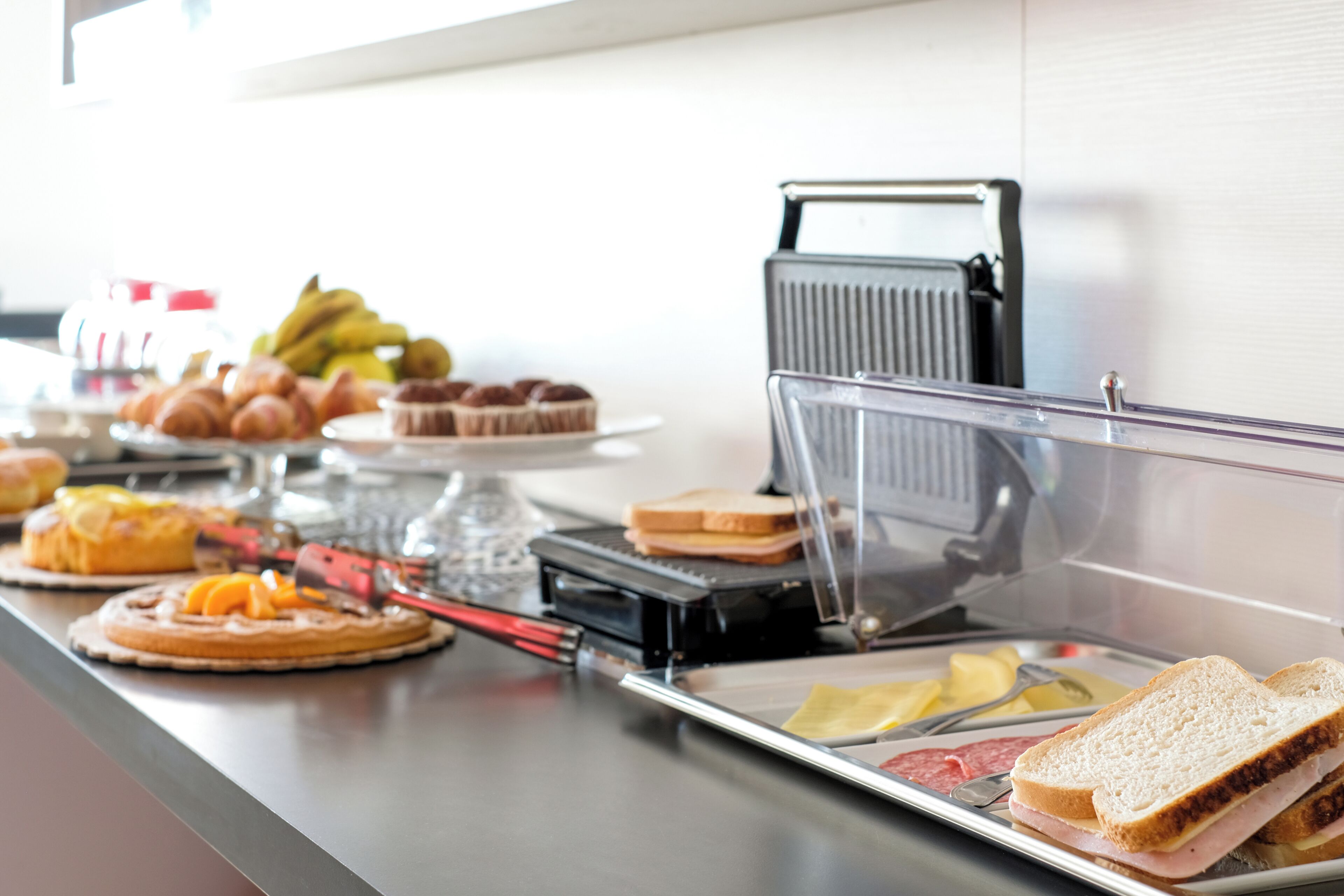 A buffet counter with various food items including pastries, bread, and a sandwich grill.