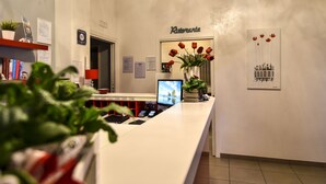 Zona de recepción de hotel con un mostrador blanco, una computadora, un florero con flores rojas y un letrero que dice 