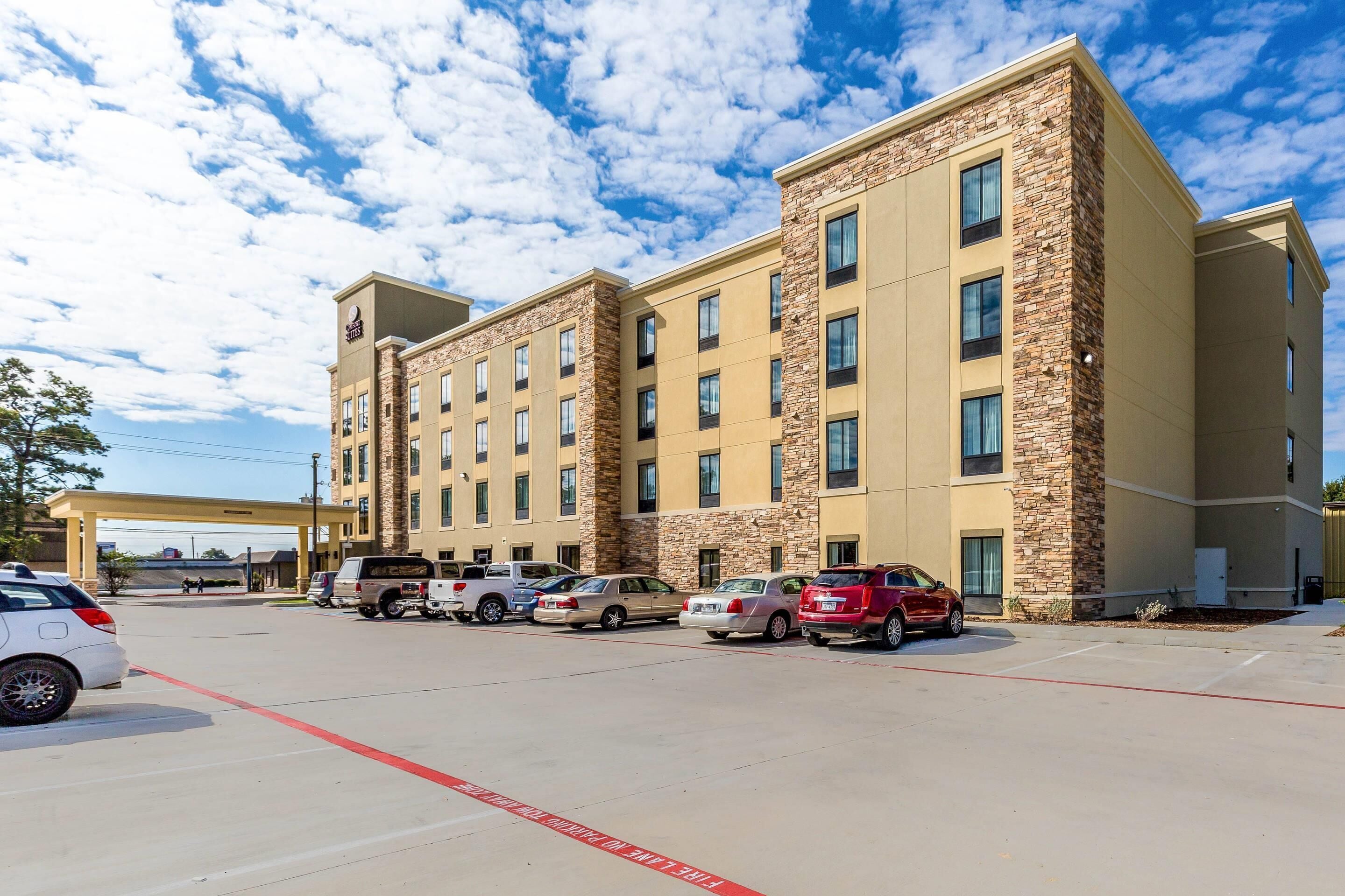 A multi-story hotel with a stone facade, parking lot, and a clear blue sky.
