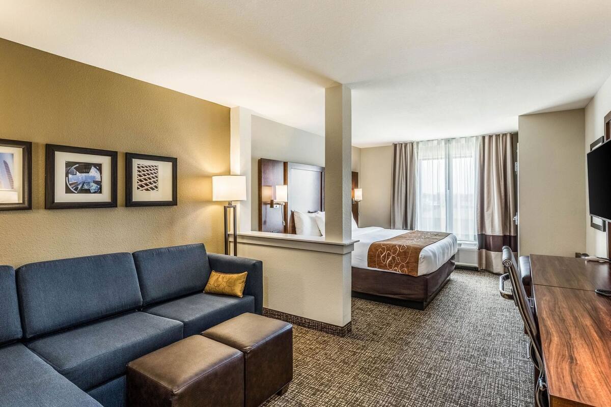 A hotel room with a sofa, ottoman, bed, TV, and a mirror.