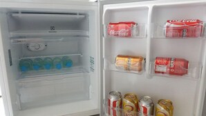 Un réfrigérateur ouvert, dont les étagères contiennent des bouteilles d’eau, des canettes de Coca-Cola et des canettes de SKOL.