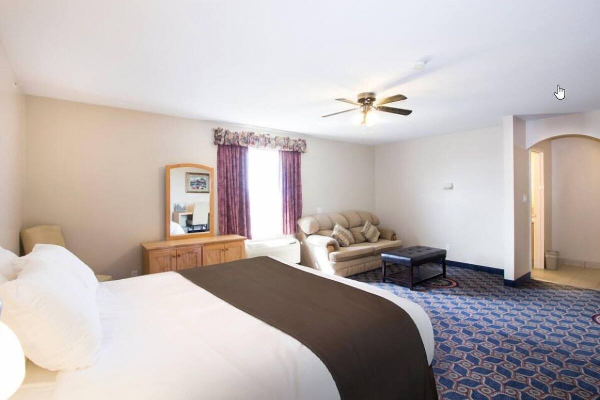A hotel room with a bed, a sofa, a small table, a ceiling fan, and a door leading to another room.