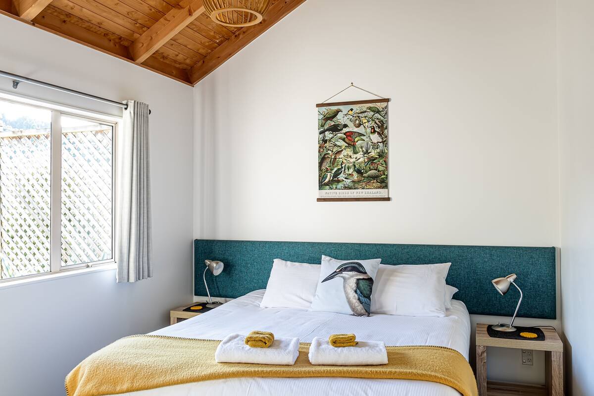 A hotel room with a bed, two nightstands, a wall-mounted poster of New Zealand native birds, and a window with curtains.