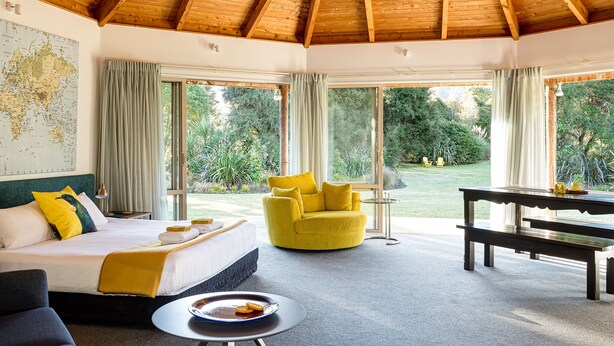 A room with a wooden ceiling, a bed with a map on the wall, a yellow armchair, a dining table, and large windows with curtains. - Abel Tasman Lodge (Marahau)
