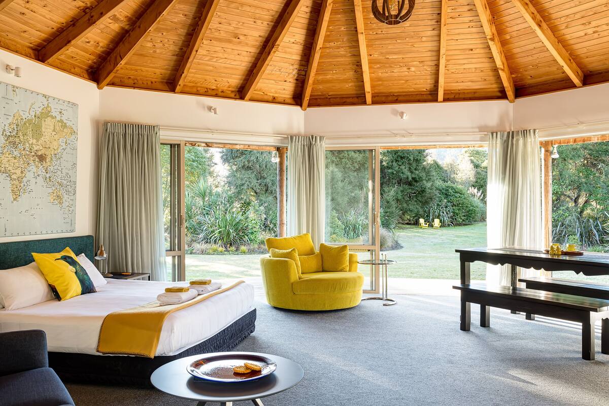 A room with a wooden ceiling, a bed with a map on the wall, a yellow armchair, a dining table, and large windows with curtains.