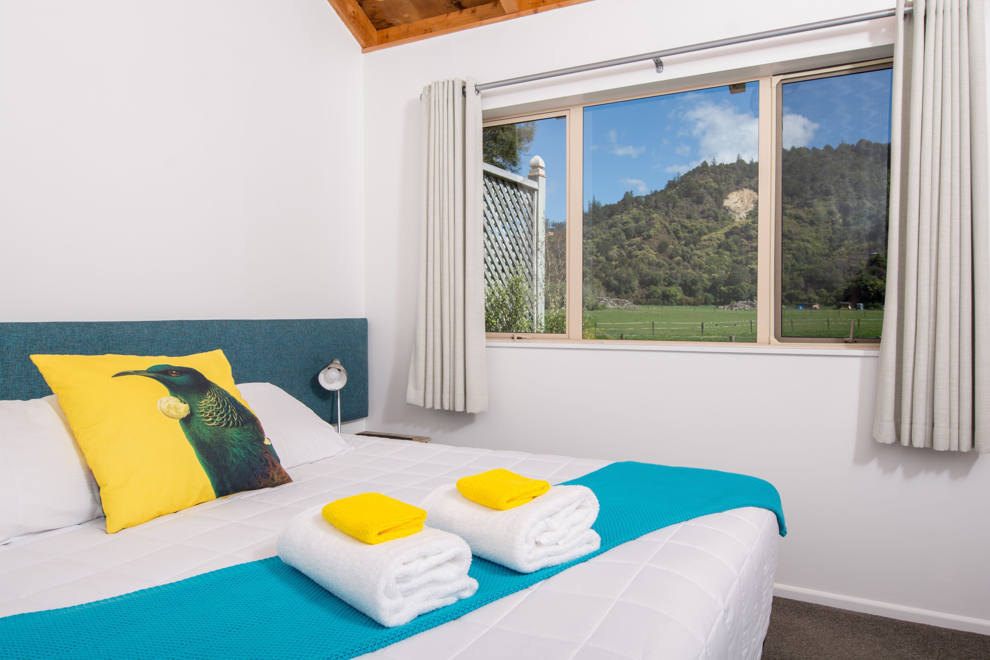 Un dormitorio con cama, dos toallas, un almohadón, una ventana con vistas a un bosque y una cortina.