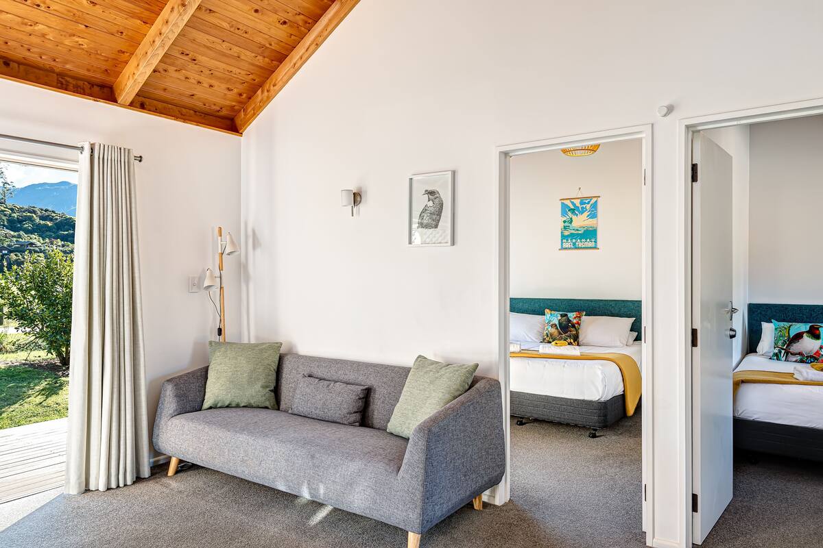 A modern room with a grey sofa, a bed with colorful bedding, a wooden ceiling, and a view of greenery outside.