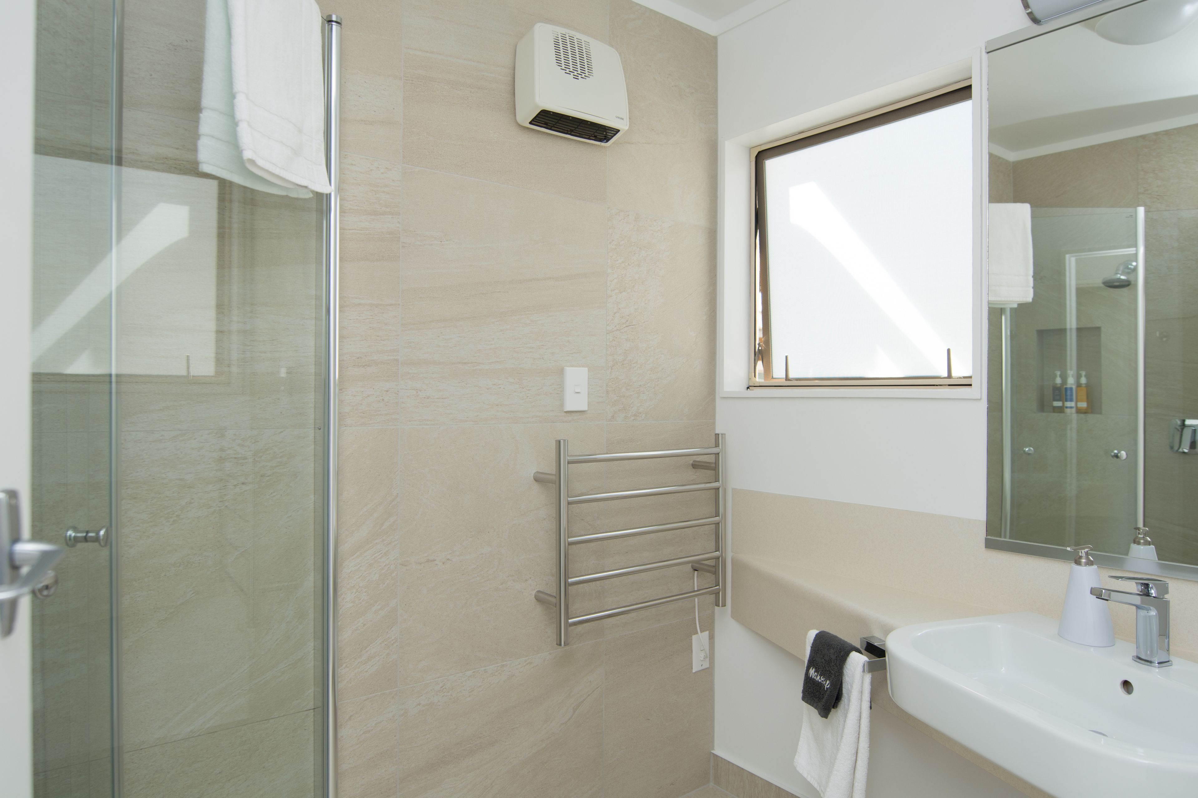 A modern bathroom with a glass shower enclosure, a towel rack, a wall-mounted air conditioner, and a large mirror above a sink.