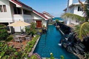 Piscina con cascada, rodeada de exuberante follaje y un patio con mobiliario exterior.