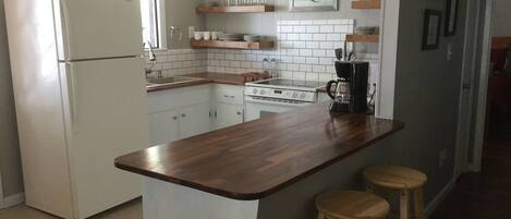 Eine Küche mit weißen Schränken, einem weißen Kühlschrank, einer Holz Arbeitsplatte, einer Kaffeemaschine und zwei Barhocker.