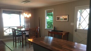 Una cocina con una isla de madera, taburetes de bar, una mesa de comedor con sillas, un ventanal con vistas a la vegetación y una puerta con un panel de vidrio con diseño de diamante.