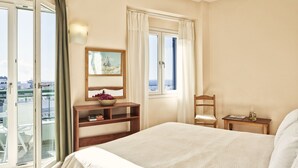 A hotel room with a bed, a wooden nightstand, a chair, a window with curtains, a view of the city, and a balcony with a glass door. - Favie Suzanne by GHH (Tinos)