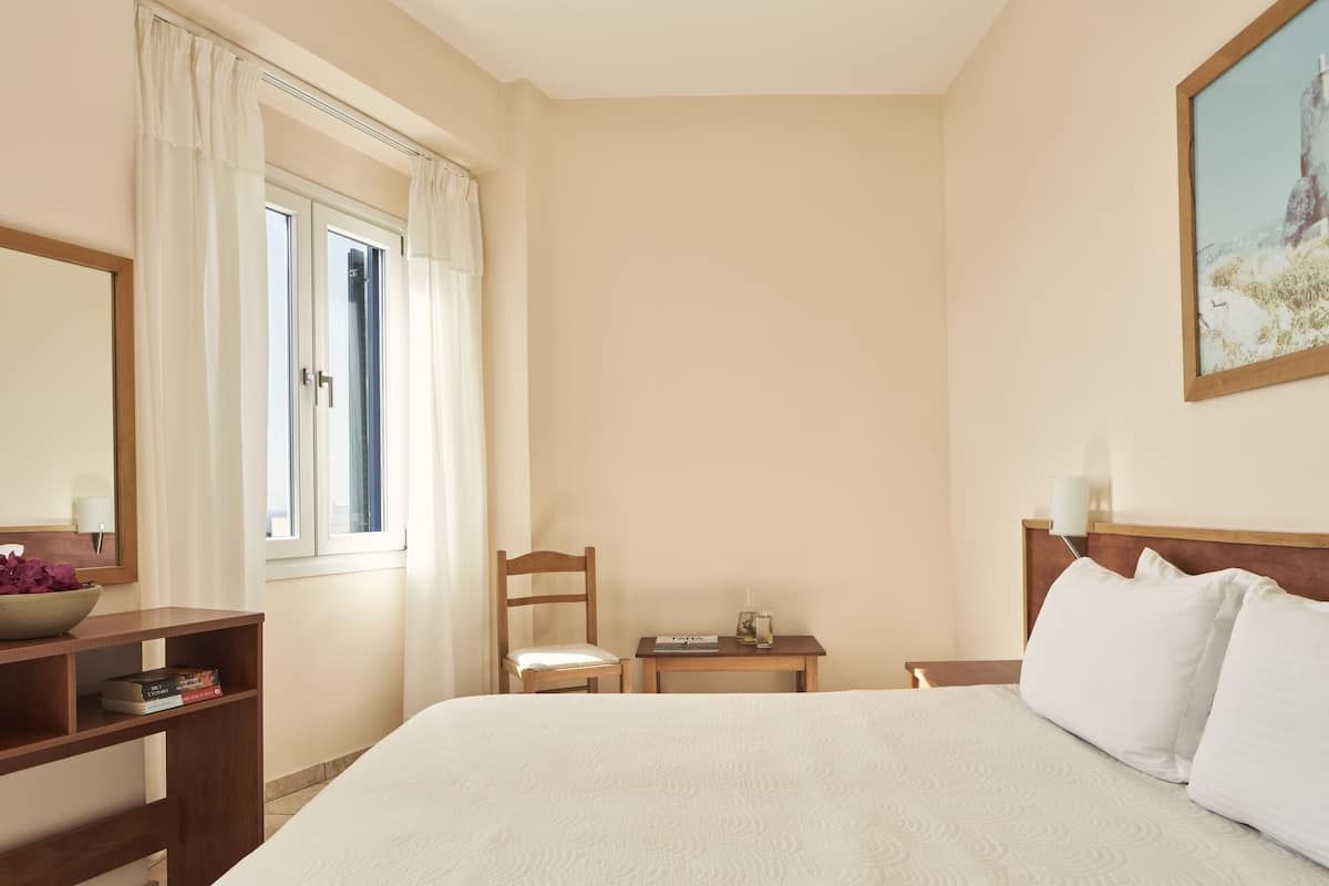 Ett hotellrum med en säng, ett träbord vid sängen, en stol, ett litet bord, ett fönster med gardiner, en tavla på väggen och ett träskrivbord med en skål och böcker.