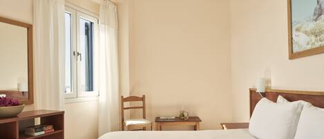 Habitación de hotel con cama, una mesita de noche de madera, una silla, una pequeña mesa, un ventanal con cortinas, un cuadro en la pared y un escritorio de madera con un cuenco y libros.