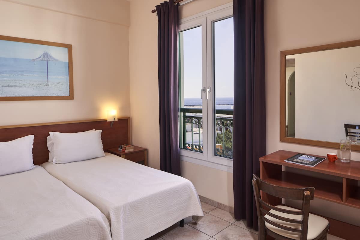 Ett hotellrum med två sängar, ett skrivbord, en stol, ett fönster med gardiner, en inramad tavla på väggen och ett litet bord med en tidning och en kopp.