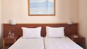 A hotel room with two beds, a wooden headboard, bedside lamps, a framed beach painting, and a small table with a glass and a bottle. - Favie Suzanne by GHH (Tinos)