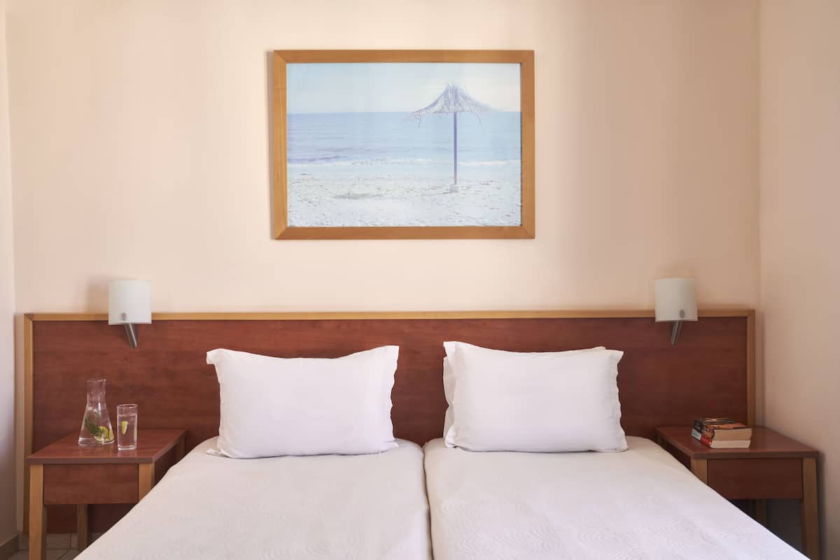 Ett hotellrum med två sängar, en sänggavel i trä, sänglampor, en inramad tavla med ett strandmotiv och ett litet bord med ett glas och en flaska.
