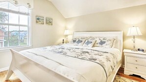 A bedroom with a bed, two bedside tables, a window, and a view of houses outside. - Comfortable and Clean Townhouse, One Block From beach (Myrtle Beach)