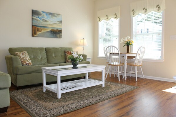 A living room with a green sofa, a white coffee table, and a dining area with white chairs and a table. - Comfortable and Clean Townhouse, One Block From beach (Myrtle Beach)
