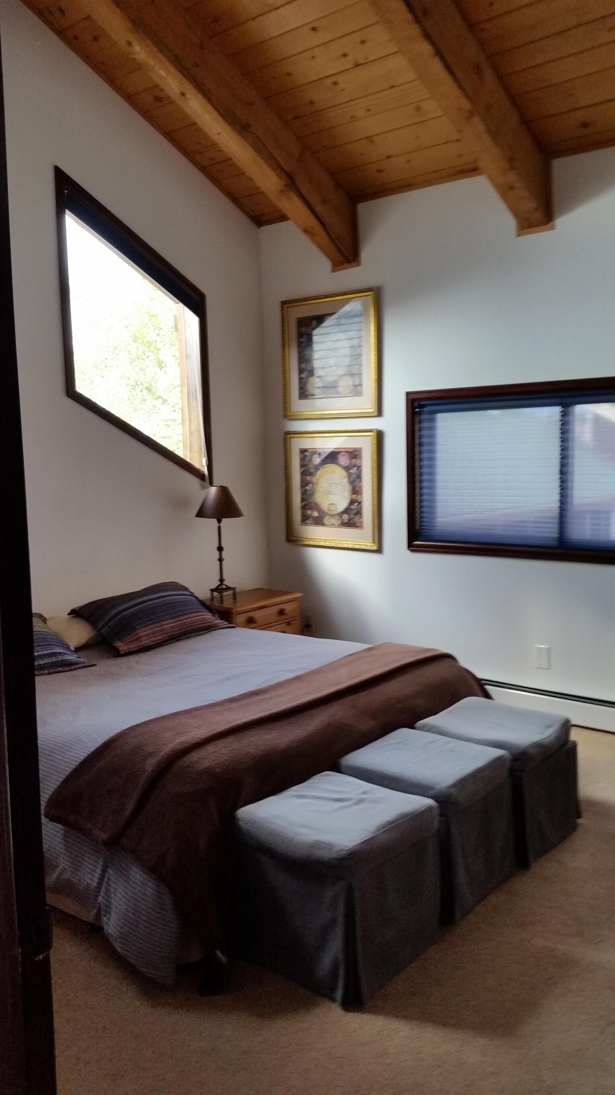Une chambre à coucher avec un lit, une table de chevet, une lampe, une fenêtre et deux tableaux encadrés accrochés au mur.