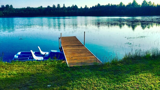 Eine Holzstege führt in einen ruhigen See, wo ein Ruderboot und ein Kajak am Ende vertäut sind.