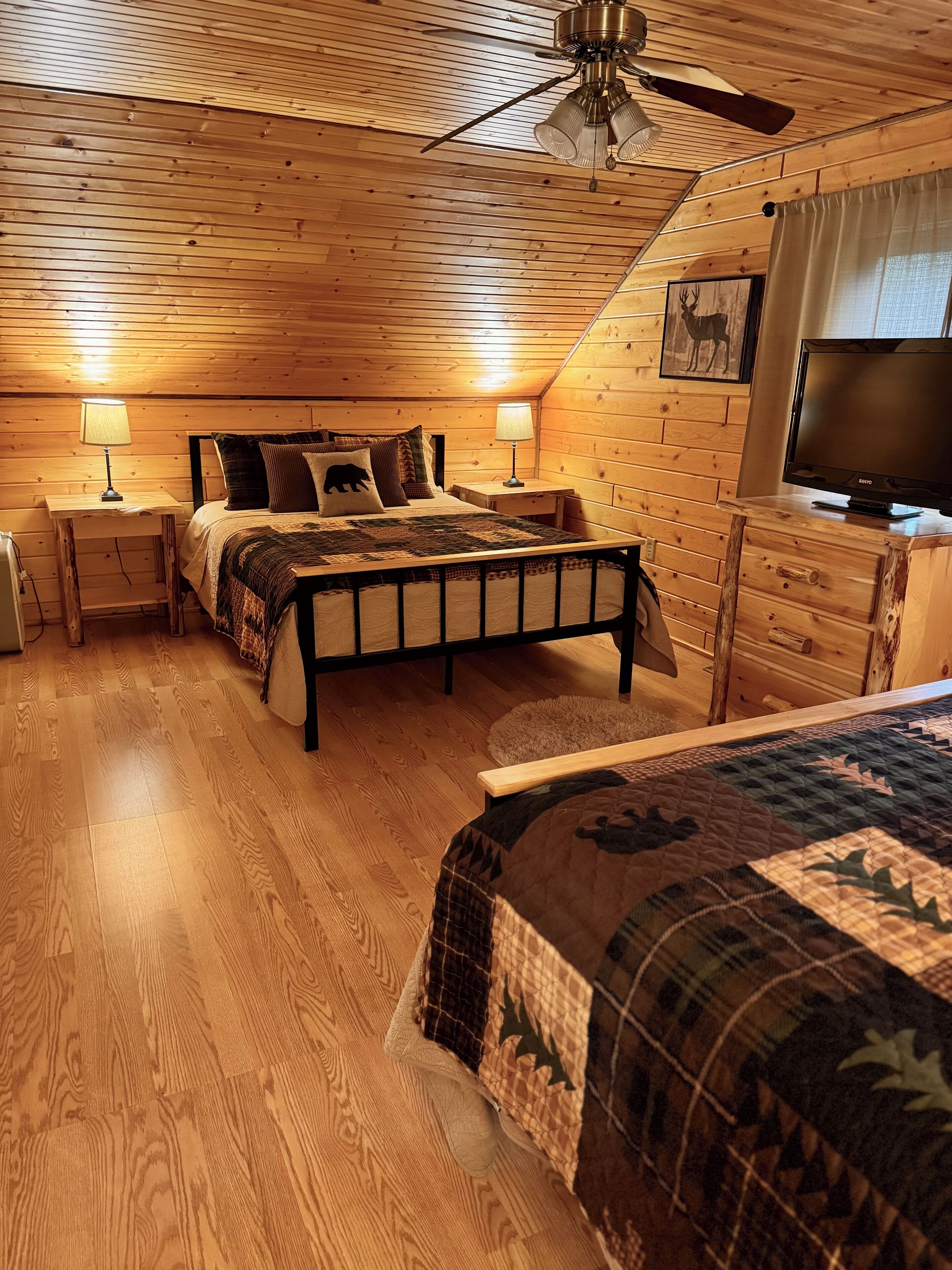Un dormitorio acogedor con techo de madera, una cama con colcha, dos lámparas de noche, un ventilador de techo y un televisor sobre un aparador de madera.