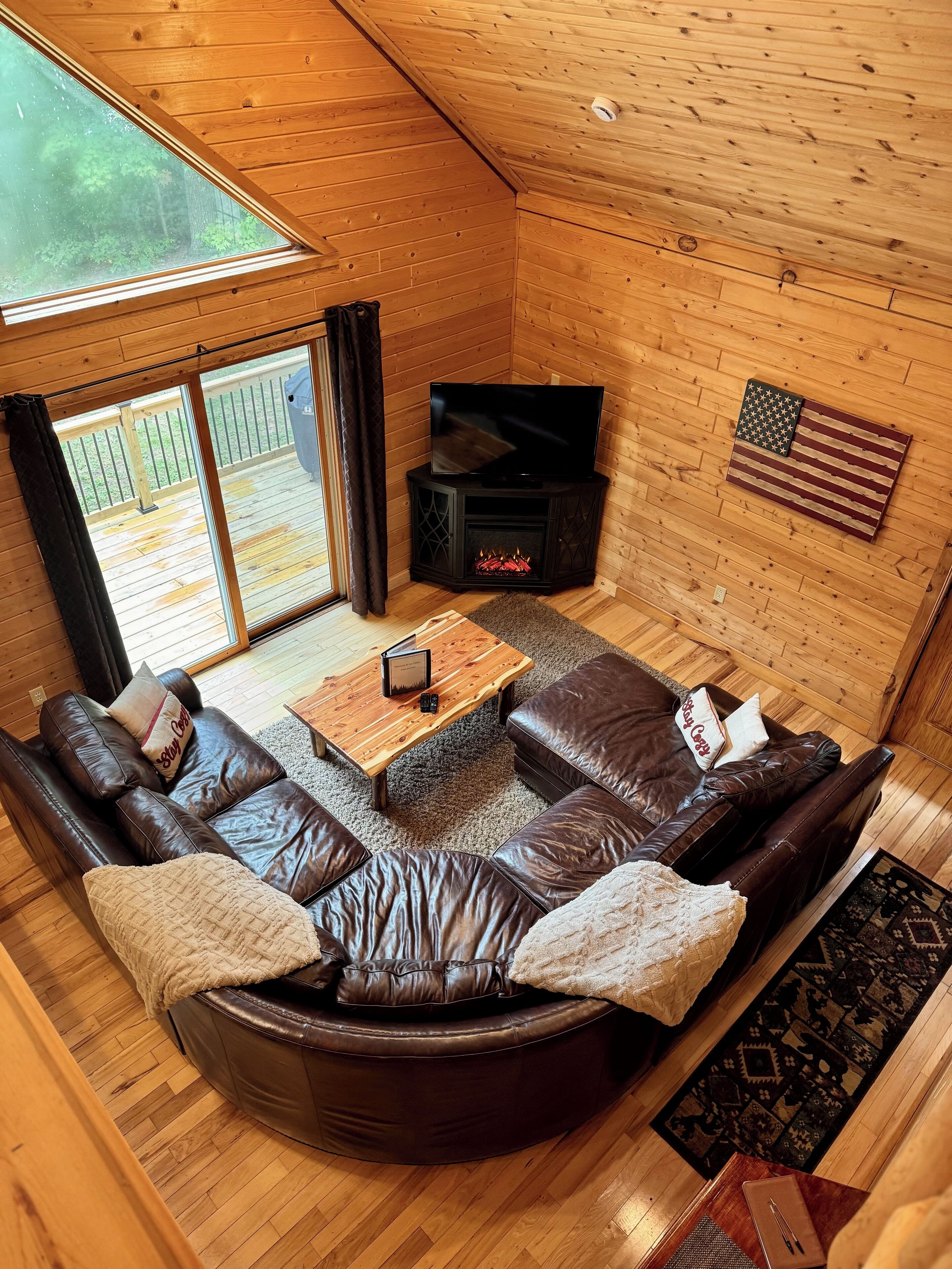 A cozy cabin interior with a leather sofa, a fireplace, a television, and a view of the outdoors.