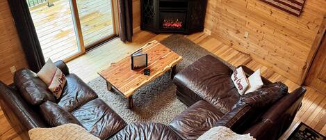 A cozy cabin interior with a leather sofa, a fireplace, a television, and a view of the outdoors.